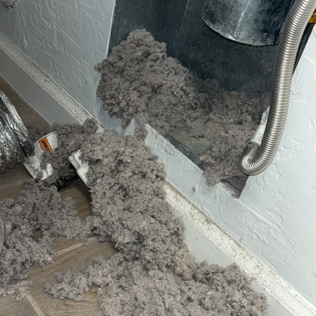 Large pile of grey dryer lint spilling from a wall vent onto a laundry room floor